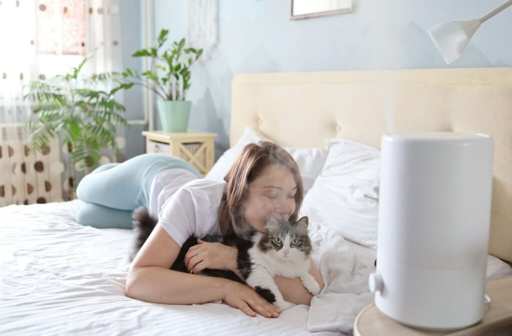 A woman cuddles her cat in bed while a humidifier blows cool mist. The bedroom is bright, with plants by the window