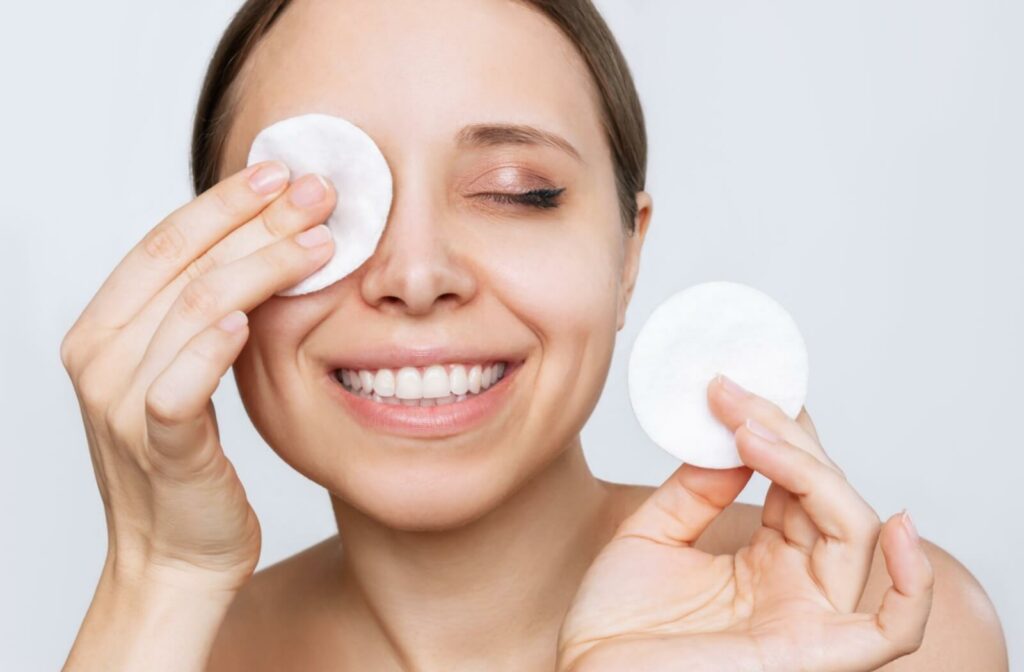 A woman holds a cotton pad in both hands, smiling while she holds one up to her eye to remove her eye makeup with.