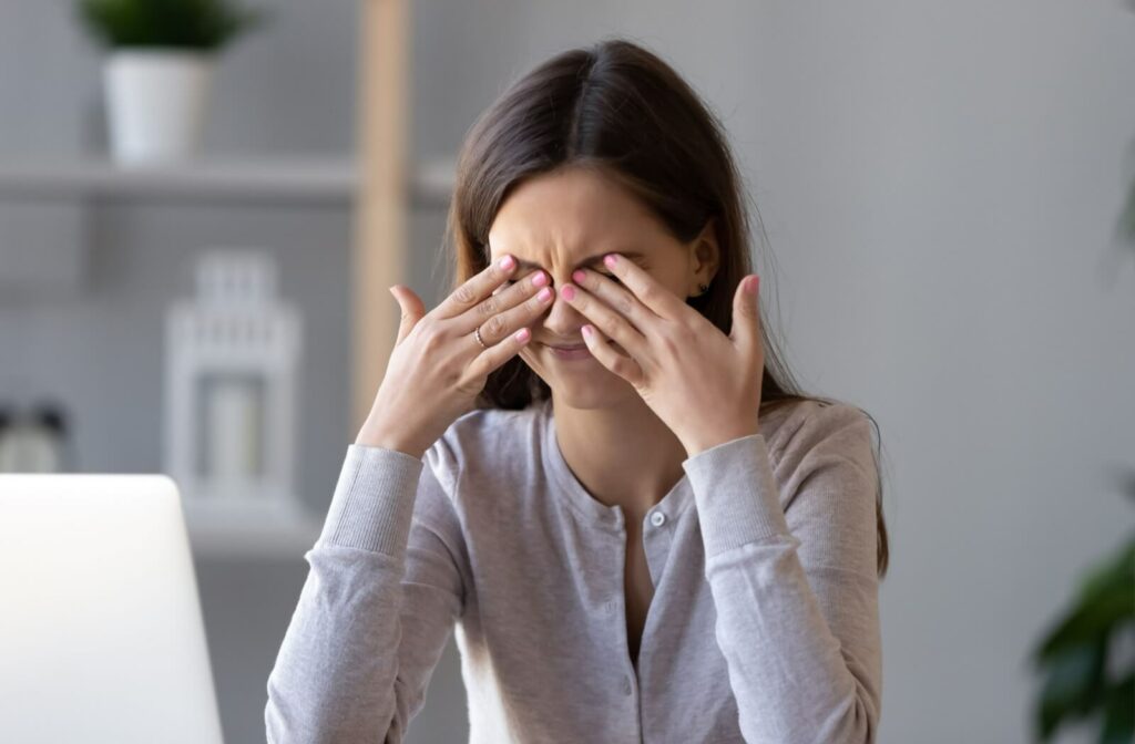 A young adult rubs their eyes with both hands, showing discomfort from dry eyes, while sitting in front of a laptop.