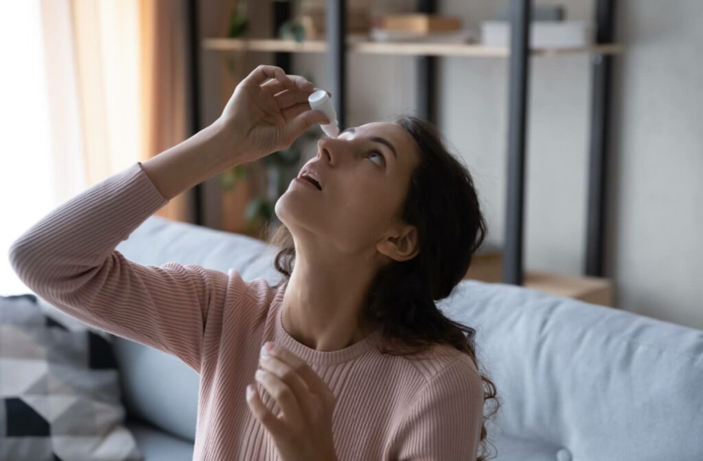 A young adult on their couch at home, tilting their head back and using specialty eye drops to find relief from their dry eyes.