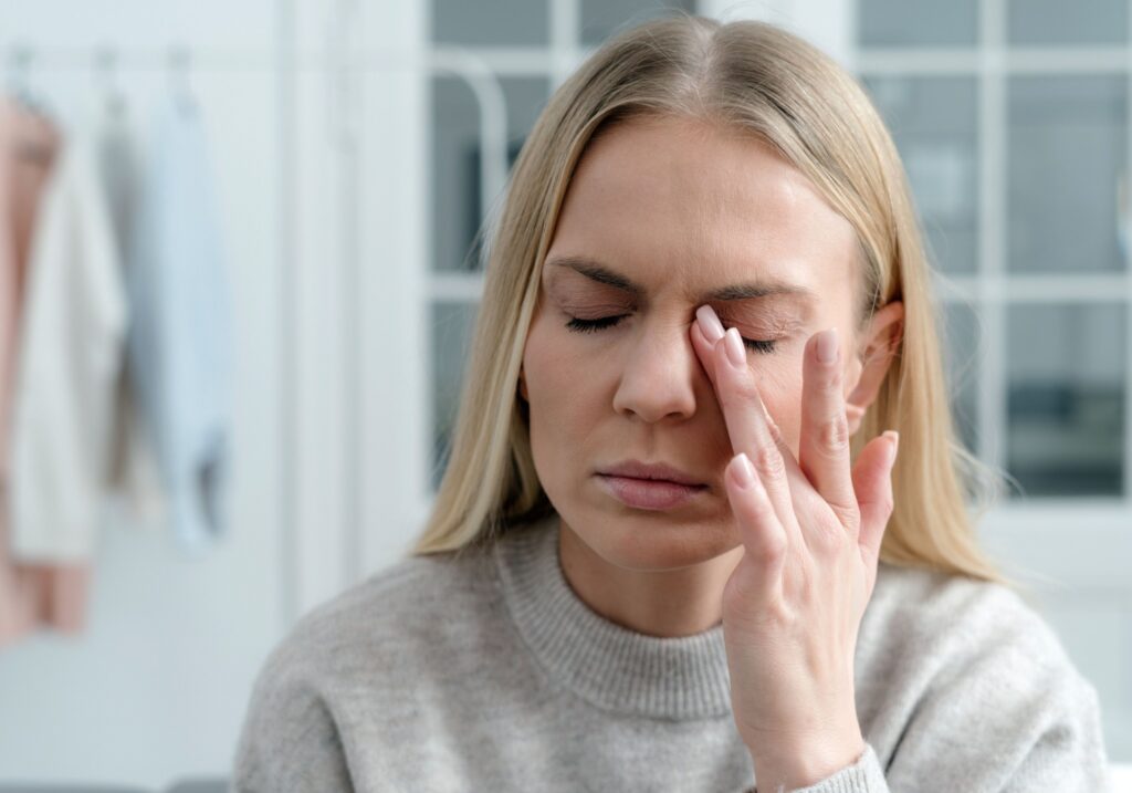 A woman with blonde hair in a grey sweater rubs her left eye with a finger.