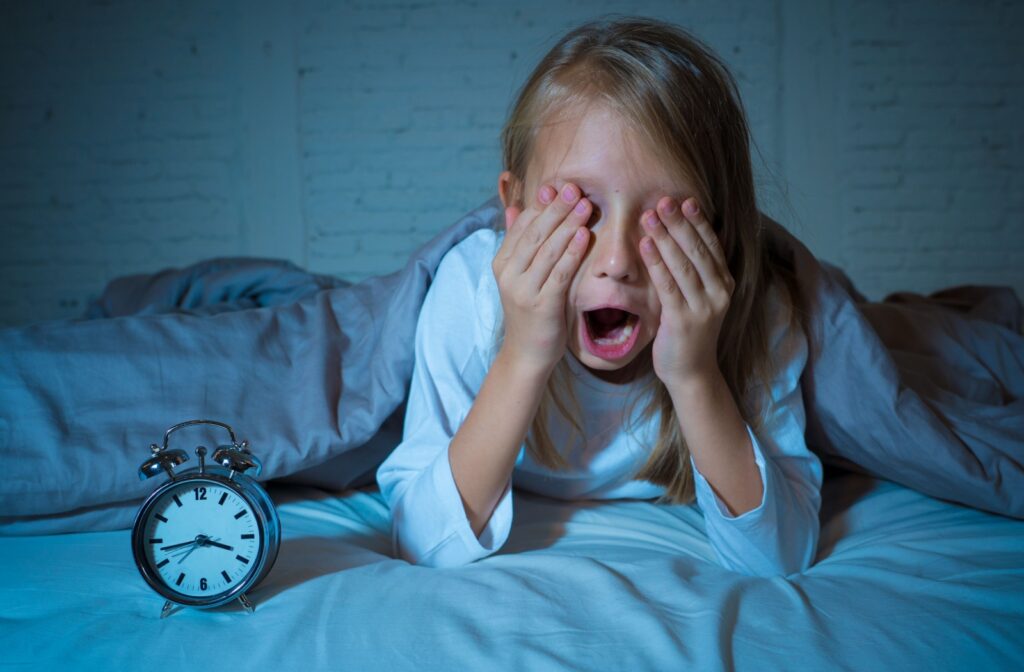 A young child yawns while rubbing their eyes in bed, an alarm clock showing that it’s almost four in the morning.