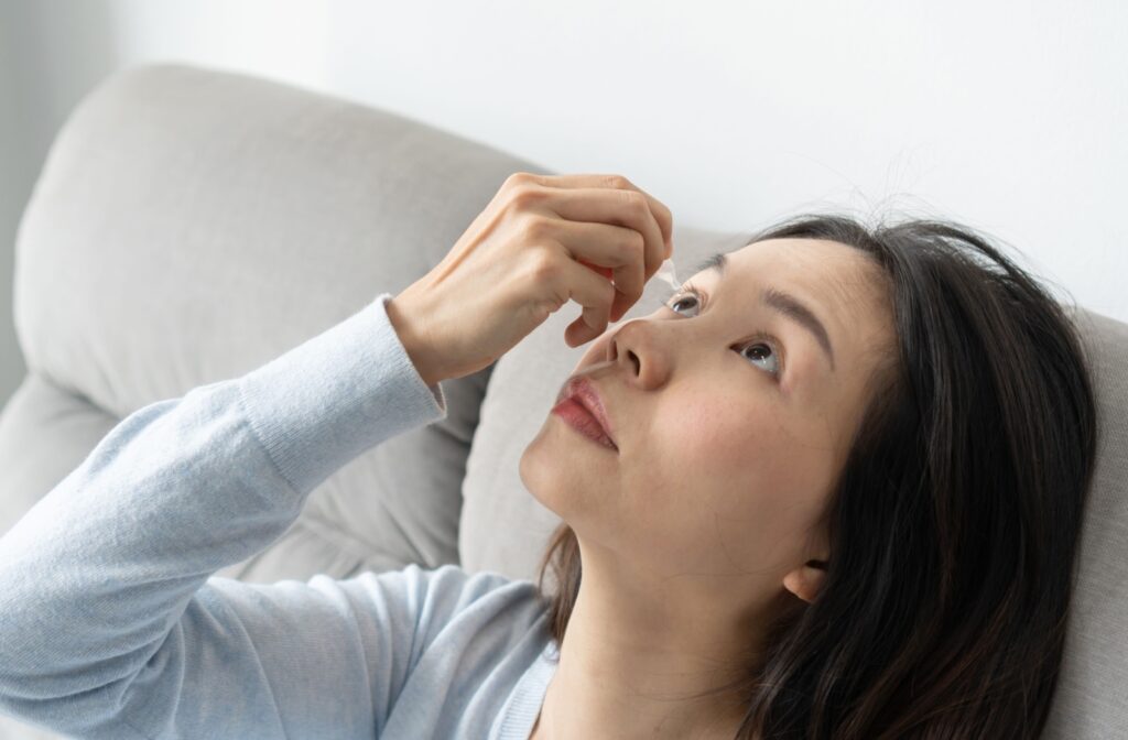 A person reclines on a couch while tilting her head back and applying eye drops to her eye.