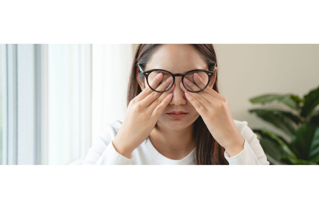 Young woman wearing glasses rubbing her eyes with both hands, showing signs of eye strain or discomfort.