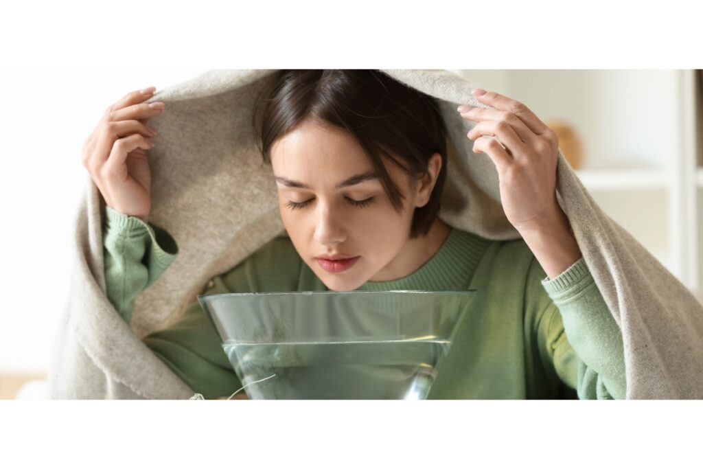 Woman sitting at a table with a towel over her head, inhaling steam from a bowl of hot water to relieve sinus congestion and pressure.